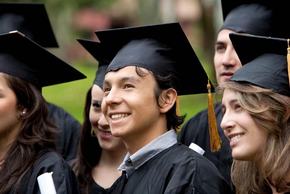 A forest green, hyperlinked, button labeled Associate Degrees in white, bolded, underlined text with an image of six graduates smile to the right of the frame in black graduate caps and gowns: A Caucasian female with sandy, brown hair stands to the right of a smiling, brown haired, Caucasian male with a blue collared shirt showing above his gown. 他的左边是一个棕色头发的人, Caucasian female stands smiling with another brown haired, Caucasian female standing partially out of the frame to her left. A Caucasian male stands partially blocked by the first female and a sixth graduate stands hidden behind the last female.